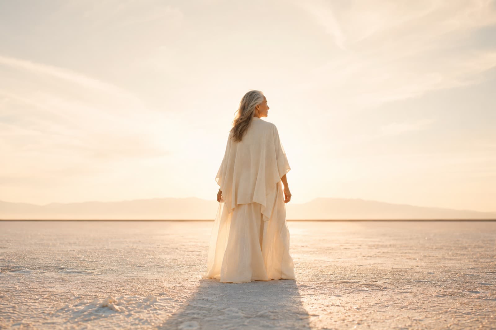 Mature woman in flowing white standing in a pale landscape surrounded by warm natural radiance.